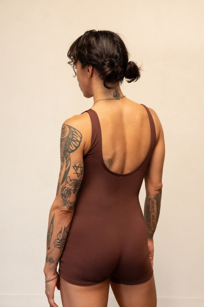 Person wearing a brown bodysuit with tattoos visible on arms and back against a beige background
