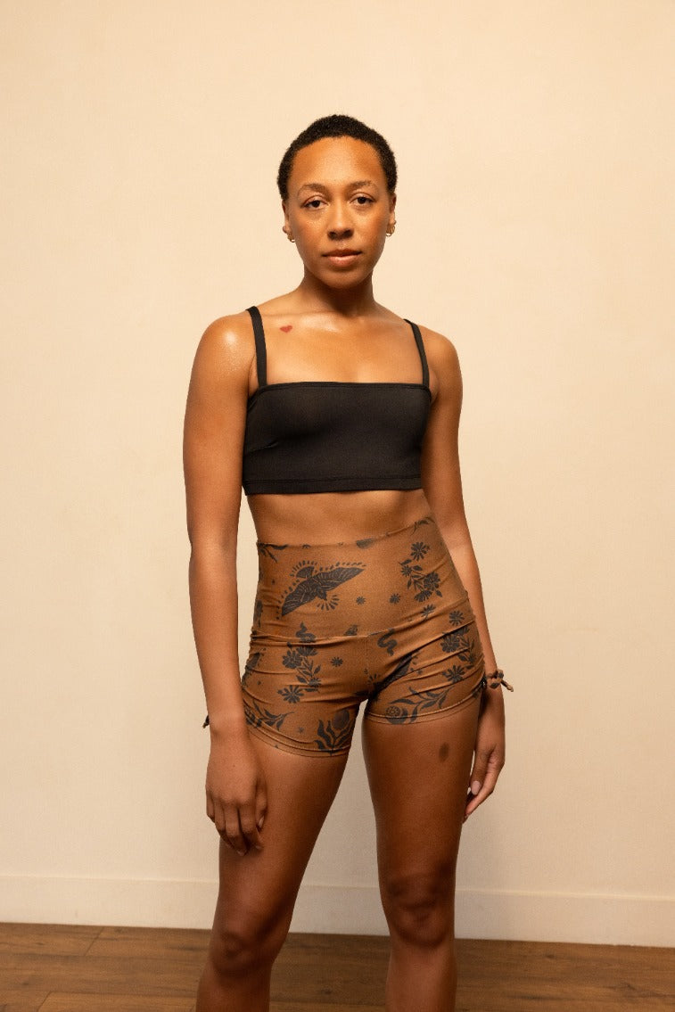 Woman wearing a black sports bra and brown patterned shorts against a beige wall.