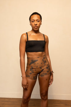 Woman wearing a black sports bra and brown patterned shorts against a beige wall.